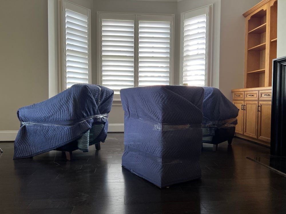 Two armchairs wrapped in blue moving blankets sit on a dark hardwood floor in a room with a bay window and cabinet.