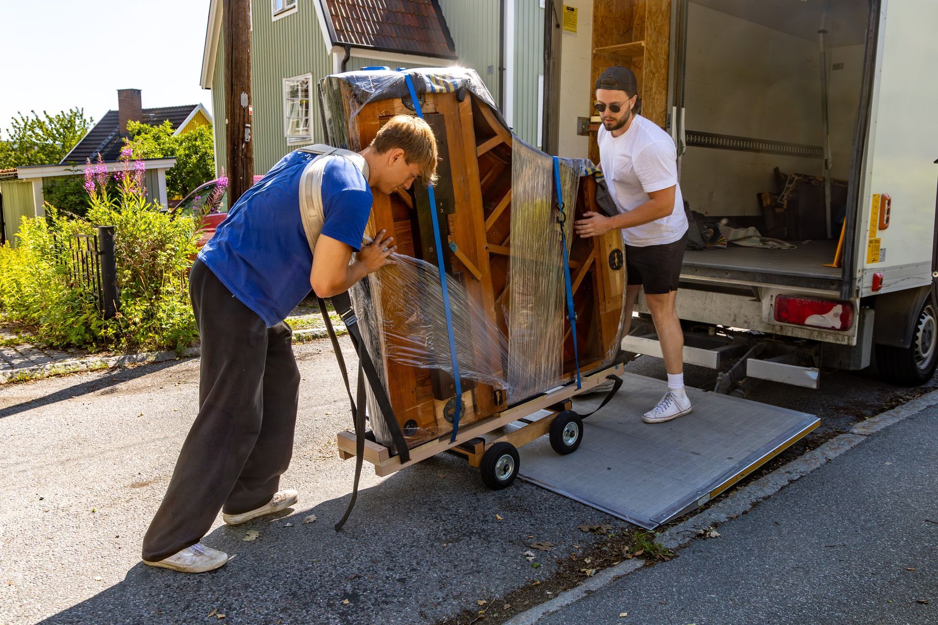 Two people loading a wrapped piano onto a truck from a dolly on a ramp.