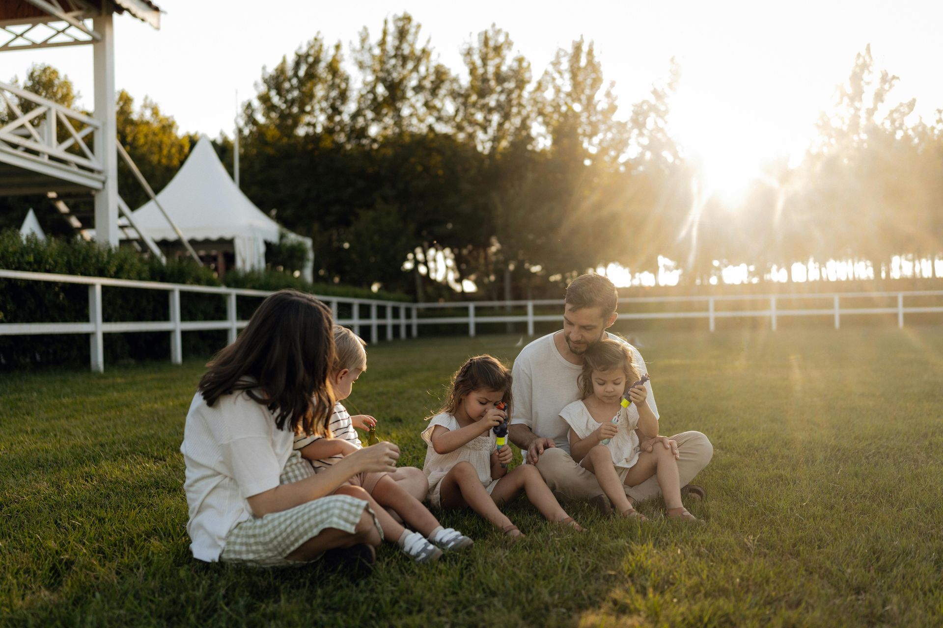 Family sitting on grass at sunset, sharing a quiet moment outdoors by a white fence and trees.