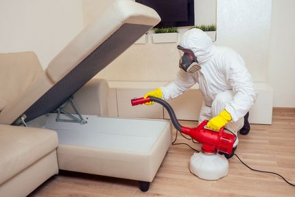 A man in a protective suit is disinfecting a couch in a living room.