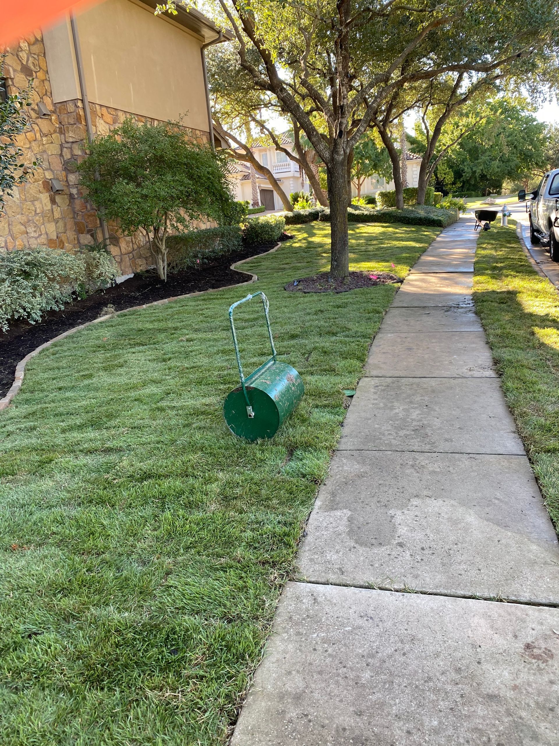 A green lawn roller is sitting on top of a lush green lawn next to a sidewalk.