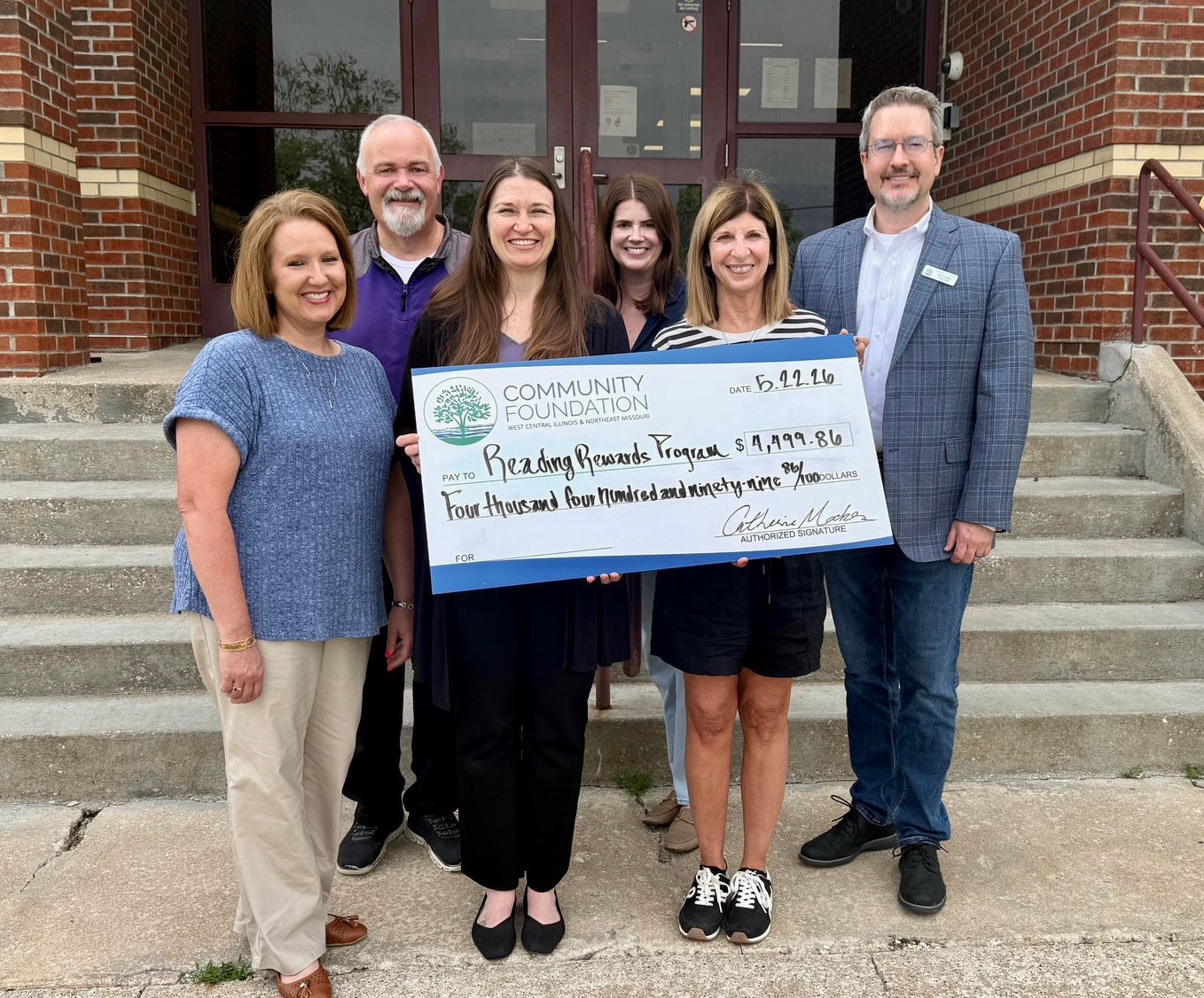 Group of people smiling and holding a donation award check in front of a school.