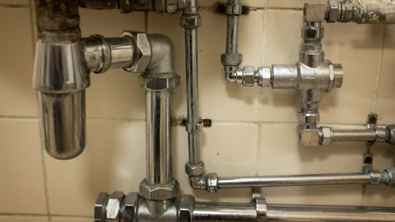 Close-up of shiny metal plumbing pipes with various connections against tiled wall.