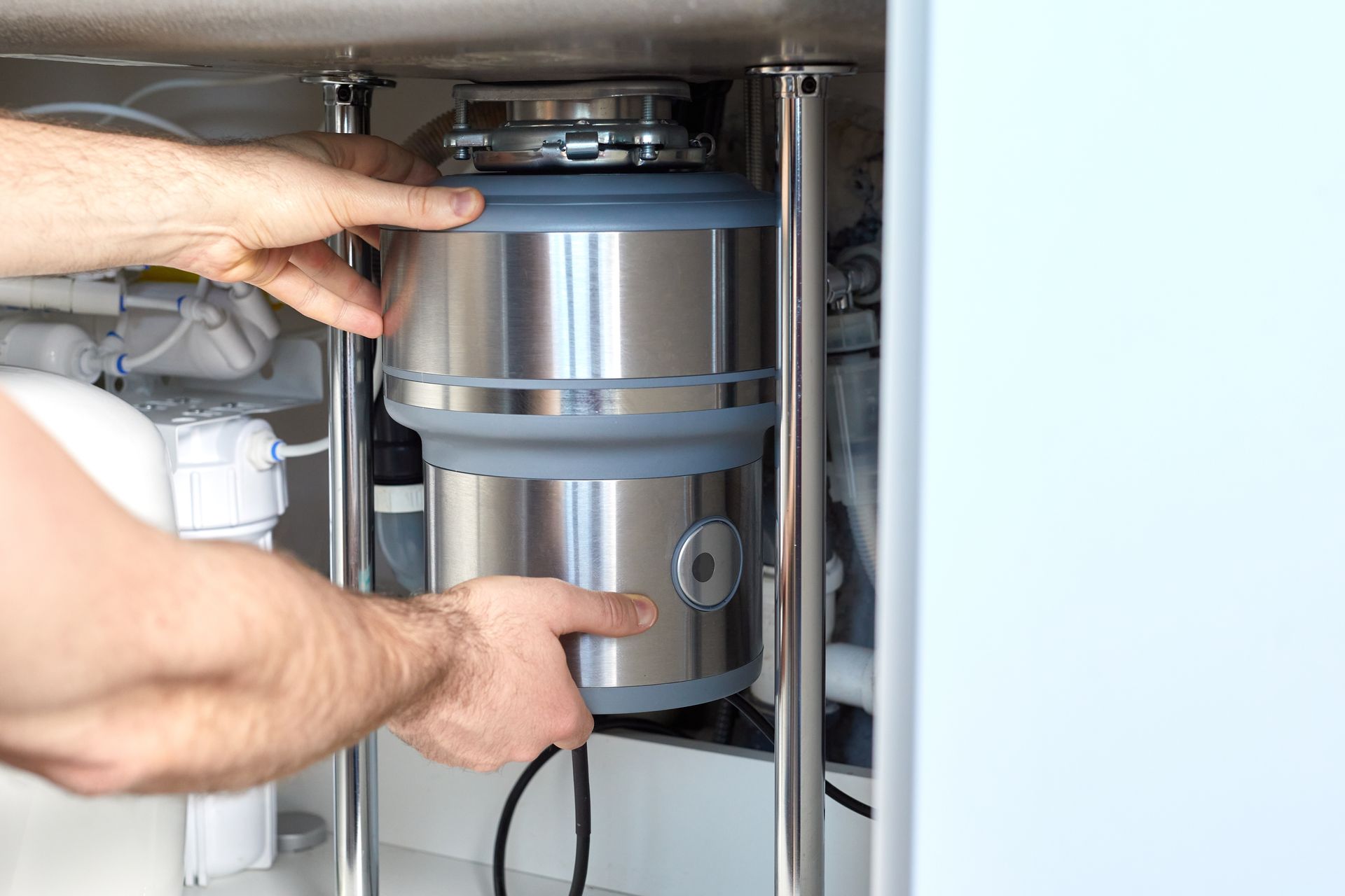 A person installing a stainless steel garbage disposal unit under a sink.