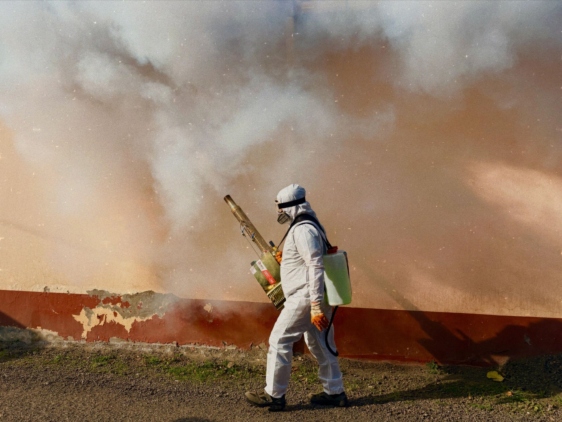 Person in protective suit spraying a wall with a fog of chemicals. Outdoors, near a building.