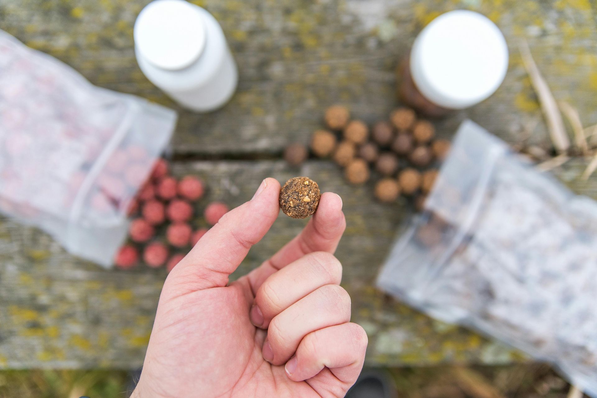 Hand holding a textured, round bait with bags of colorful bait and jars on a weathered wooden surface.