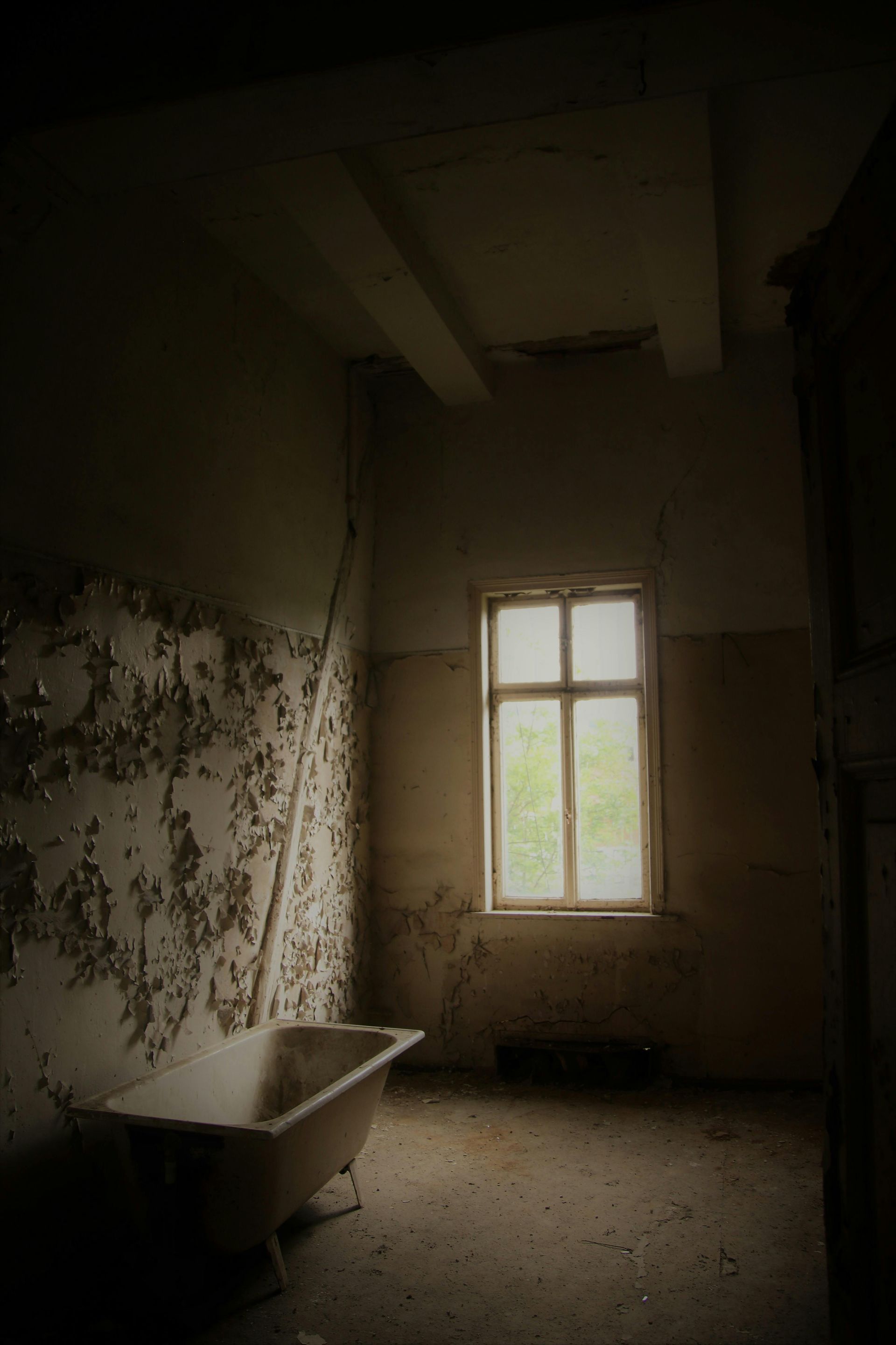 Dilapidated bathroom with a freestanding tub, peeling walls, and a window letting in light.