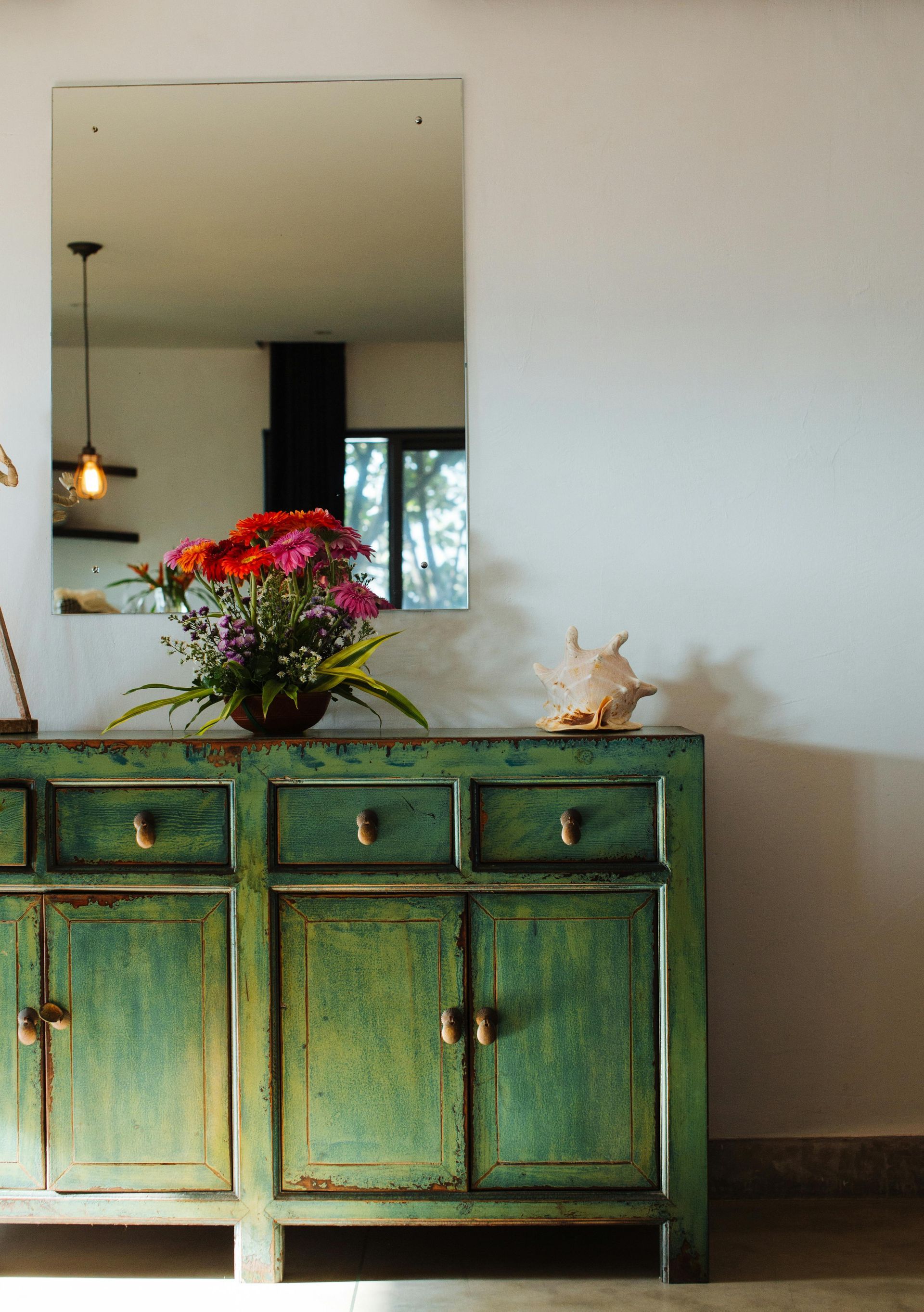 Green distressed cabinet with flowers, a seashell, and a mirror reflecting a light fixture.