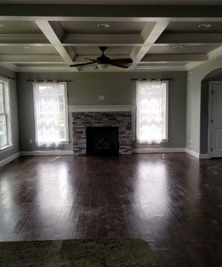 Living room with a stone fireplace, coffered ceiling, hardwood floors, and two windows with sheer curtains.