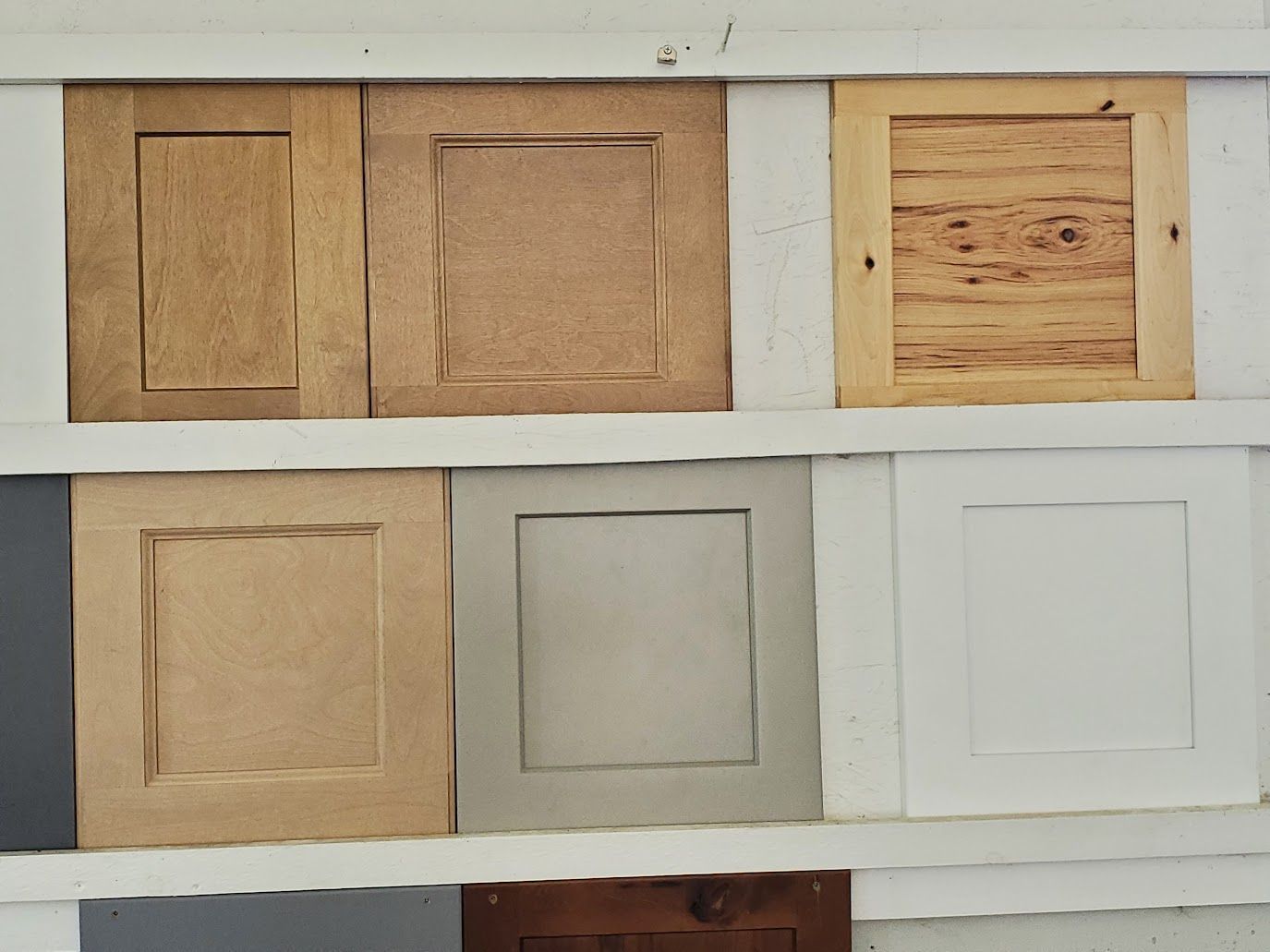 Cabinet door samples in various wood tones and painted finishes displayed on a white shelf.
