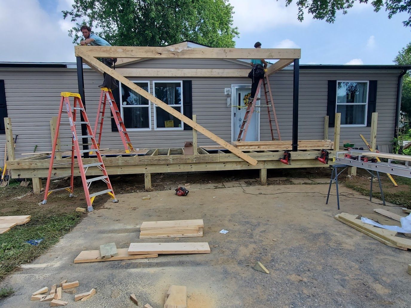Two people building a wooden porch roof on a mobile home. Ladders and lumber are present.