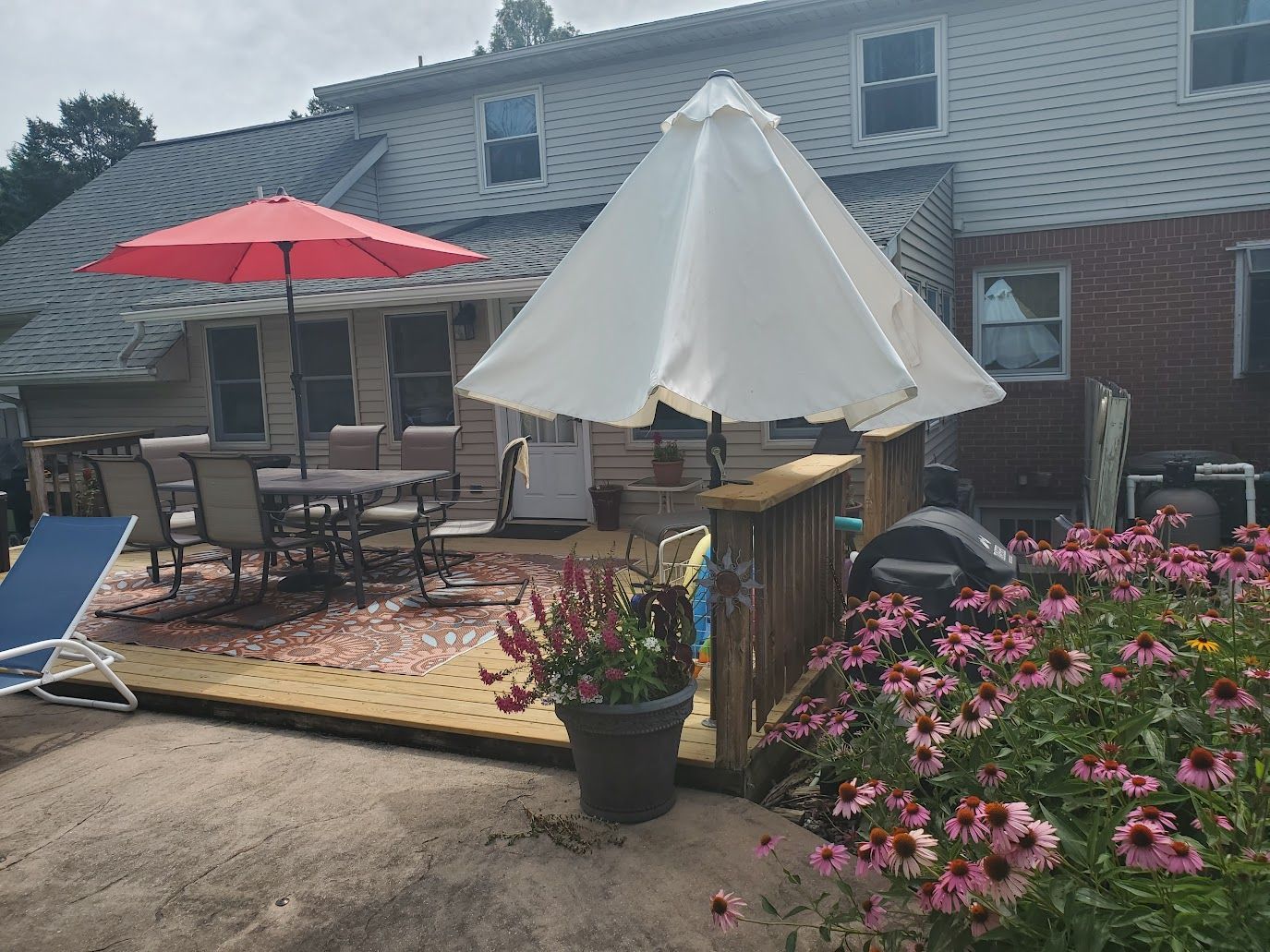 Backyard deck with table and chairs, umbrellas, and flower garden in front of a house.