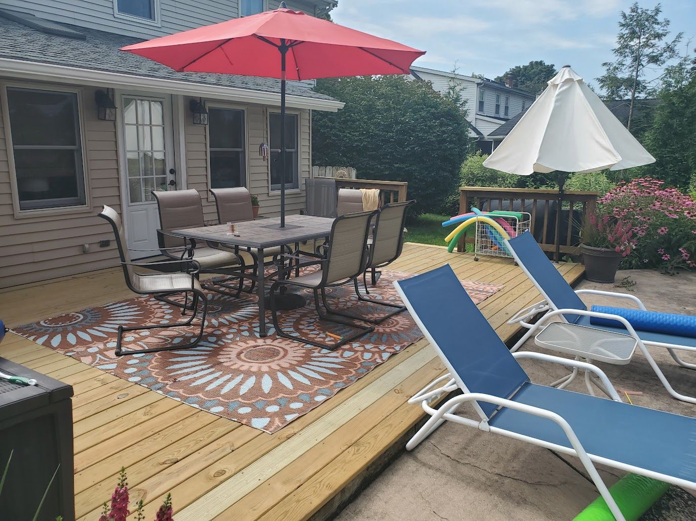 Outdoor deck with a dining table and chairs, a red umbrella, and two blue lounge chairs.