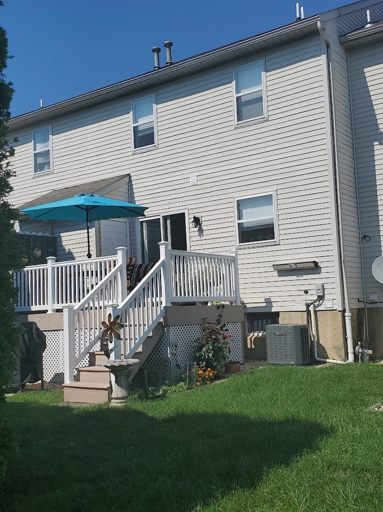 Back of house with a white deck, teal umbrella, and light-colored siding, with green grass and blue sky.