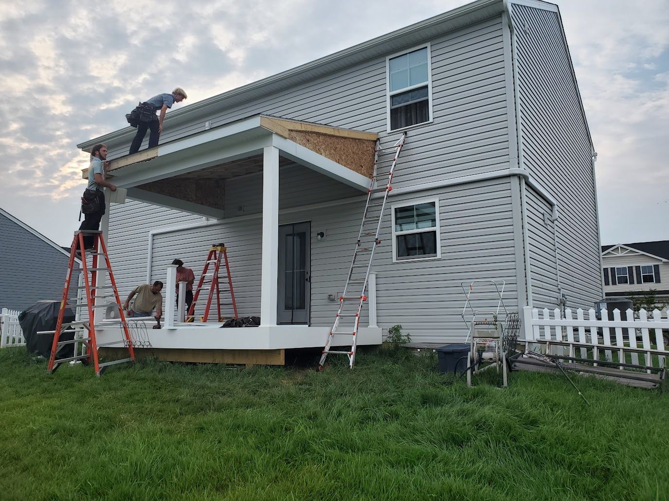 Construction workers building a covered patio on a two-story house. Ladders, tools, and materials are present.