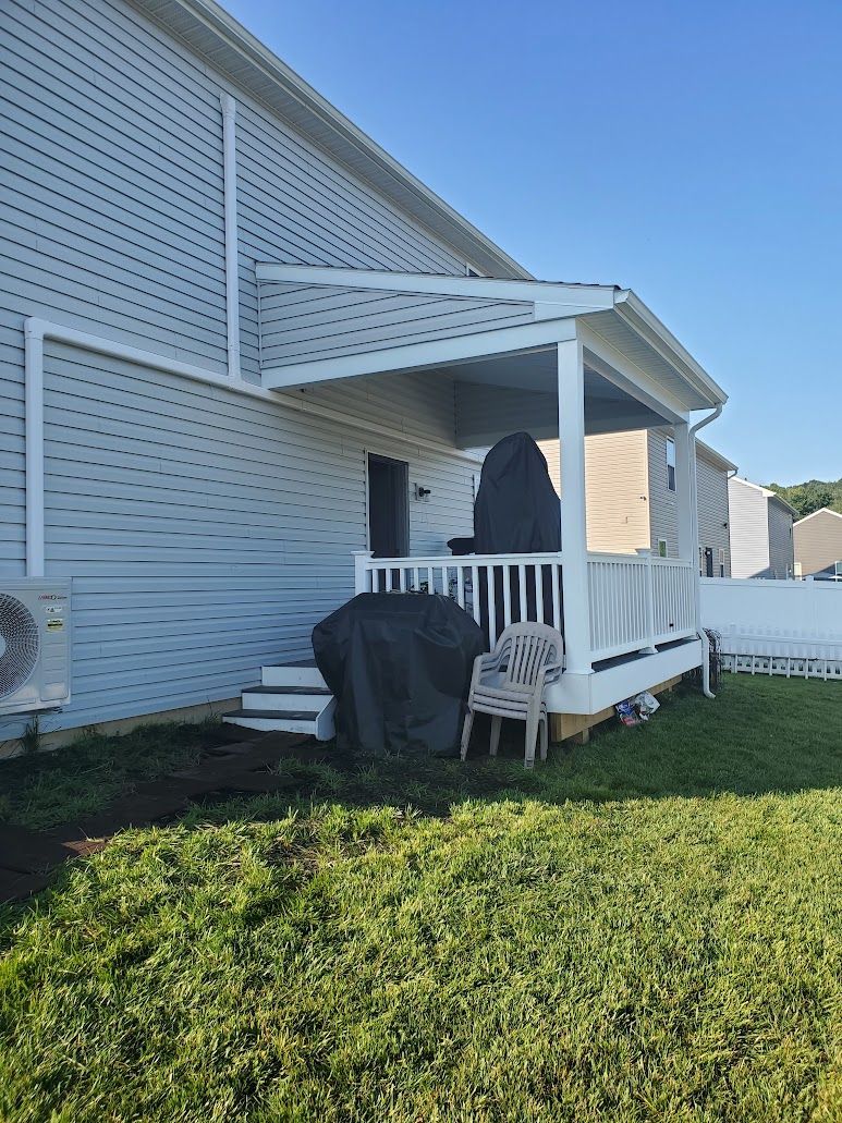 A white porch with lattice siding and a covered area, with a grill and chair on a grassy lawn.