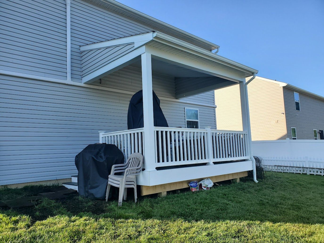 A white deck with a canopy attached to a house. Grill and a chair on the deck, grass in the foreground.