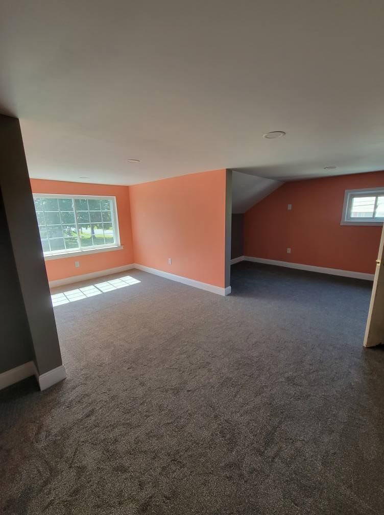 Spacious room with orange walls, gray carpet, and two windows, with a partial view of a smaller adjacent room.