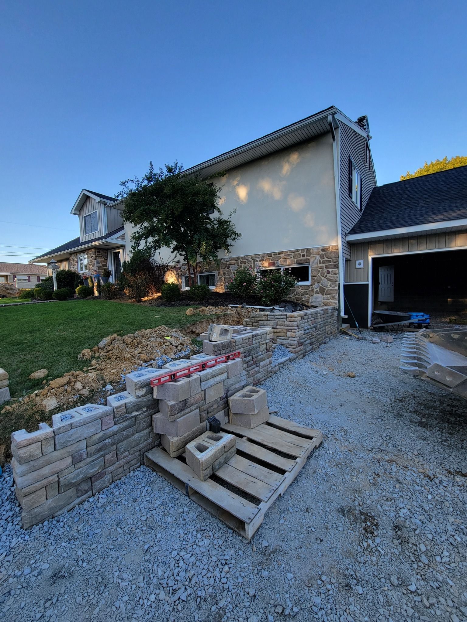 A house with a stone facade has a retaining wall being built on a gravel driveway.