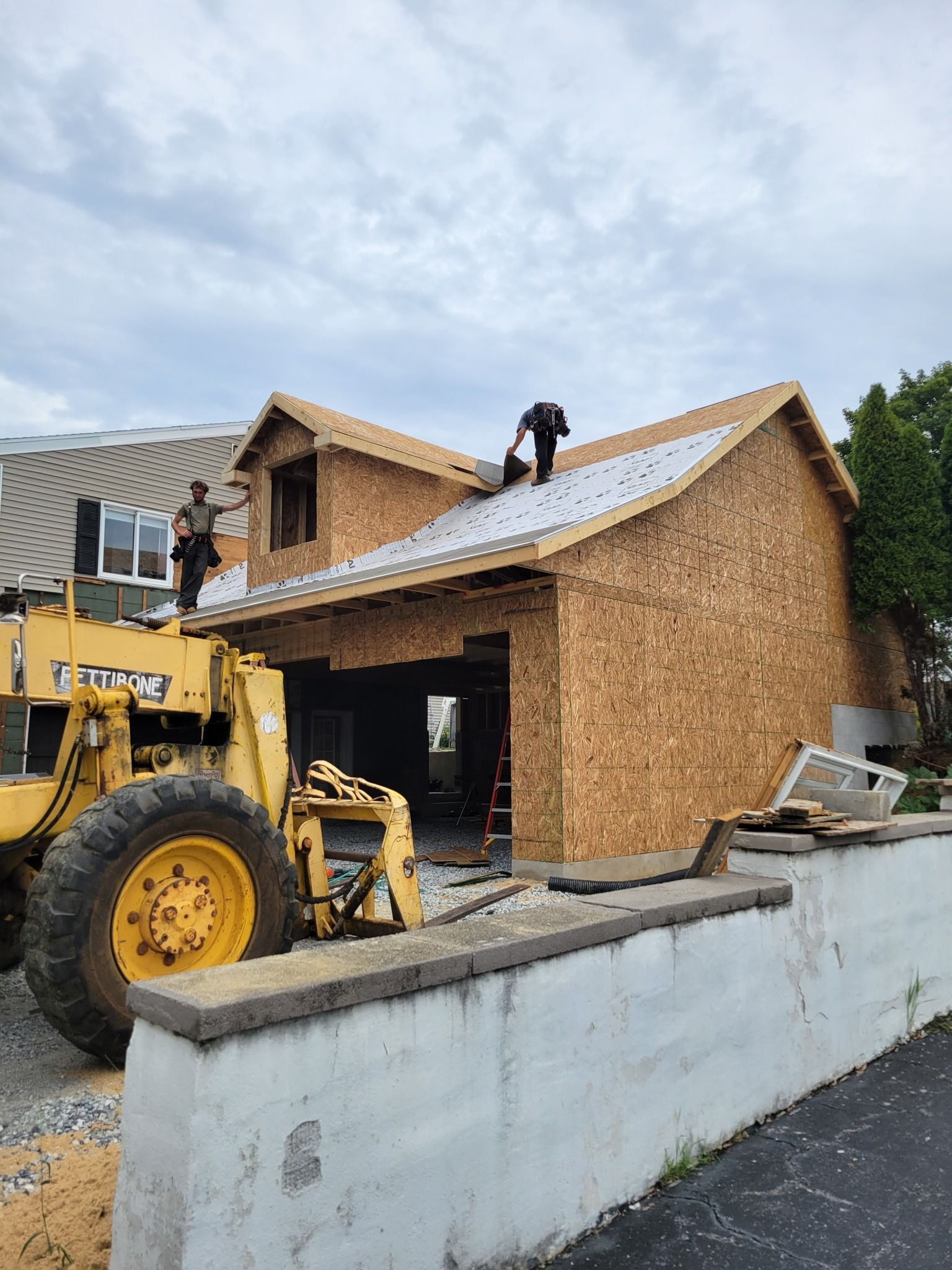 Construction workers on a roof and on a machine, building a house, under a cloudy sky.