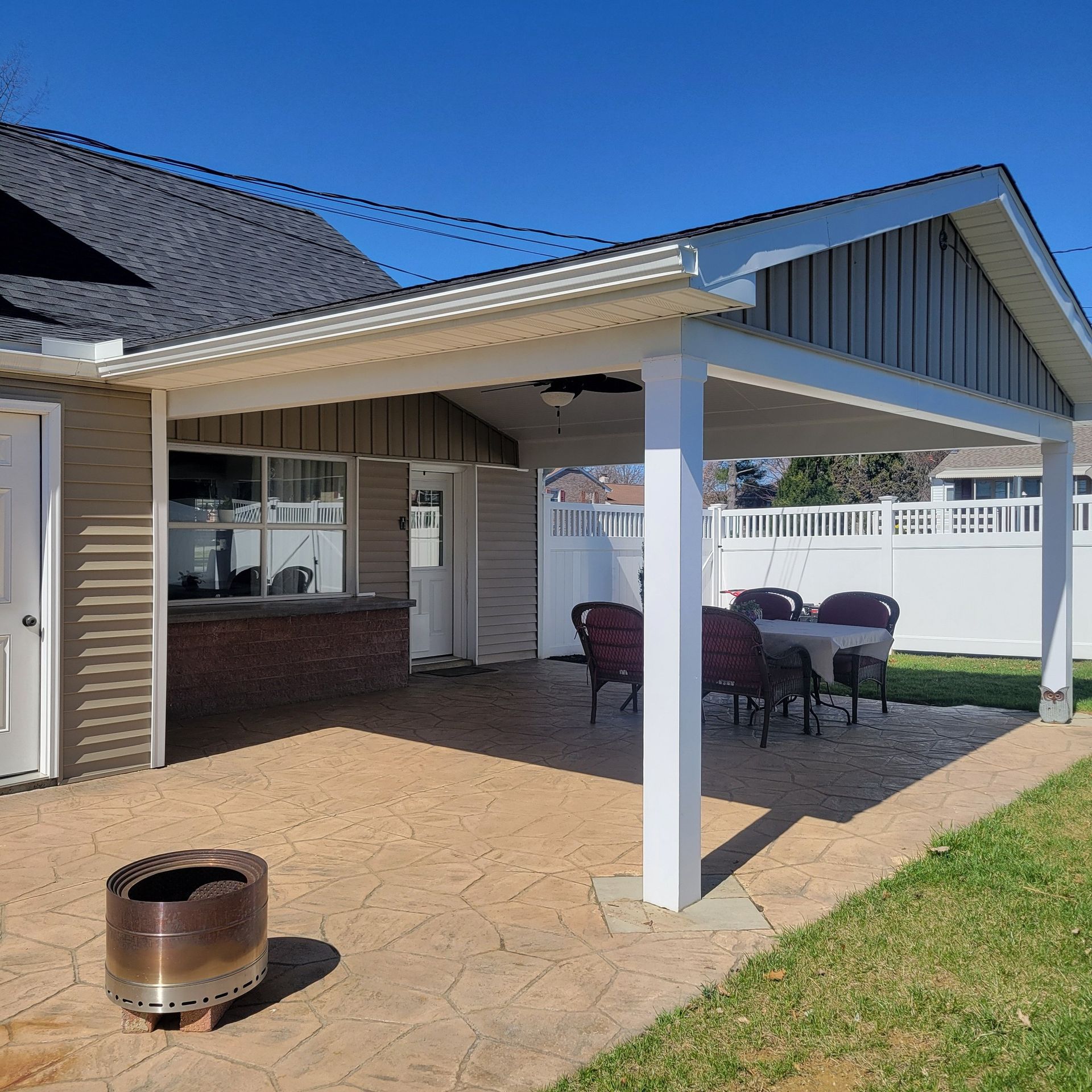 A backyard patio with an attached covered porch, a table and chairs, and a fire pit under a clear blue sky.