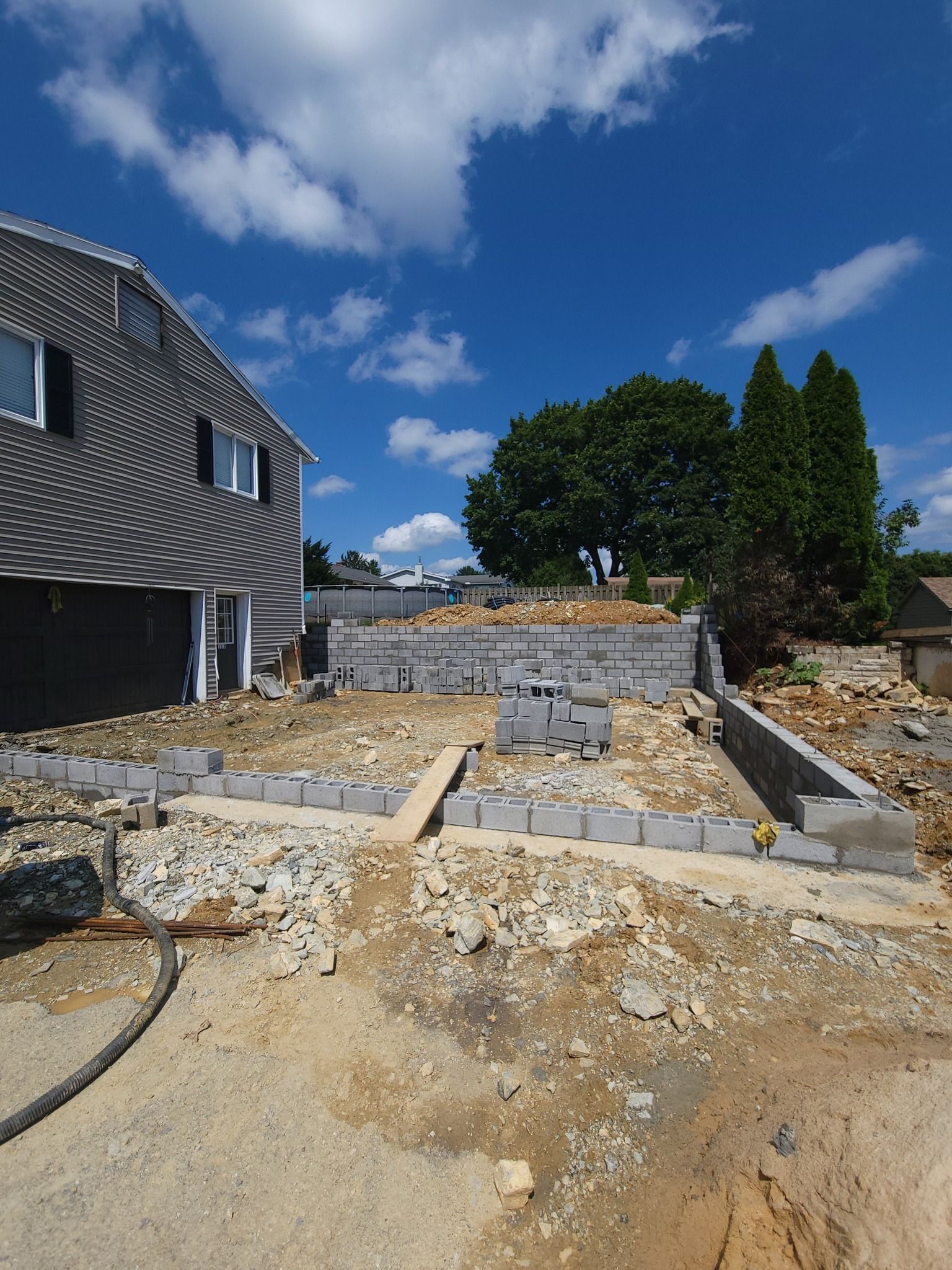 Construction site with gray concrete blocks, sunny blue sky, existing home to the left.