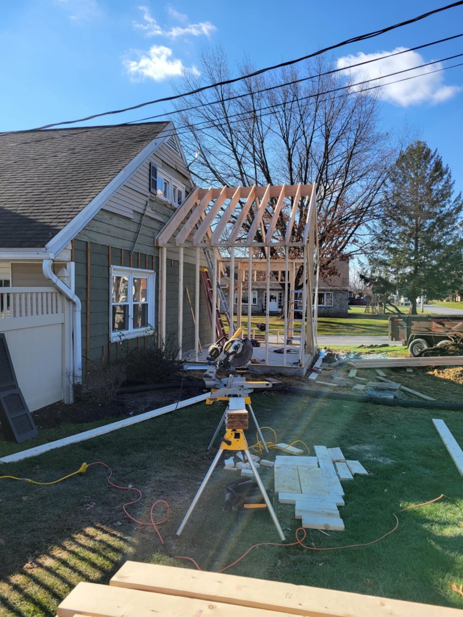 Construction of a new structure attached to a green house. Saw and lumber sit on a grassy yard under a sunny sky.