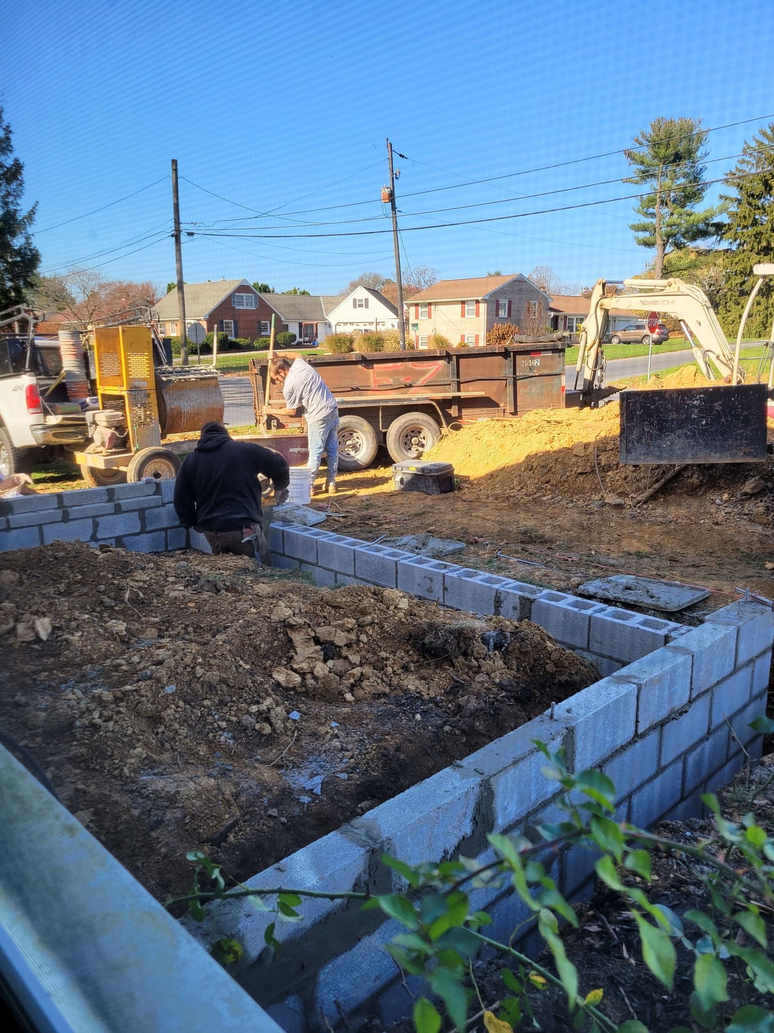Construction site: Workers laying blocks, preparing foundation. Trucks, excavator, houses in background. Sunny day.