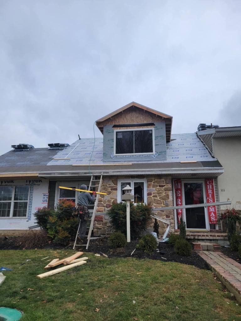 House under construction; worker on a ladder; new roof being installed. Overcast day.