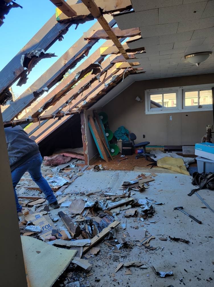 A person removing roof material in a damaged attic with debris on the floor; light streaming in from open beams.