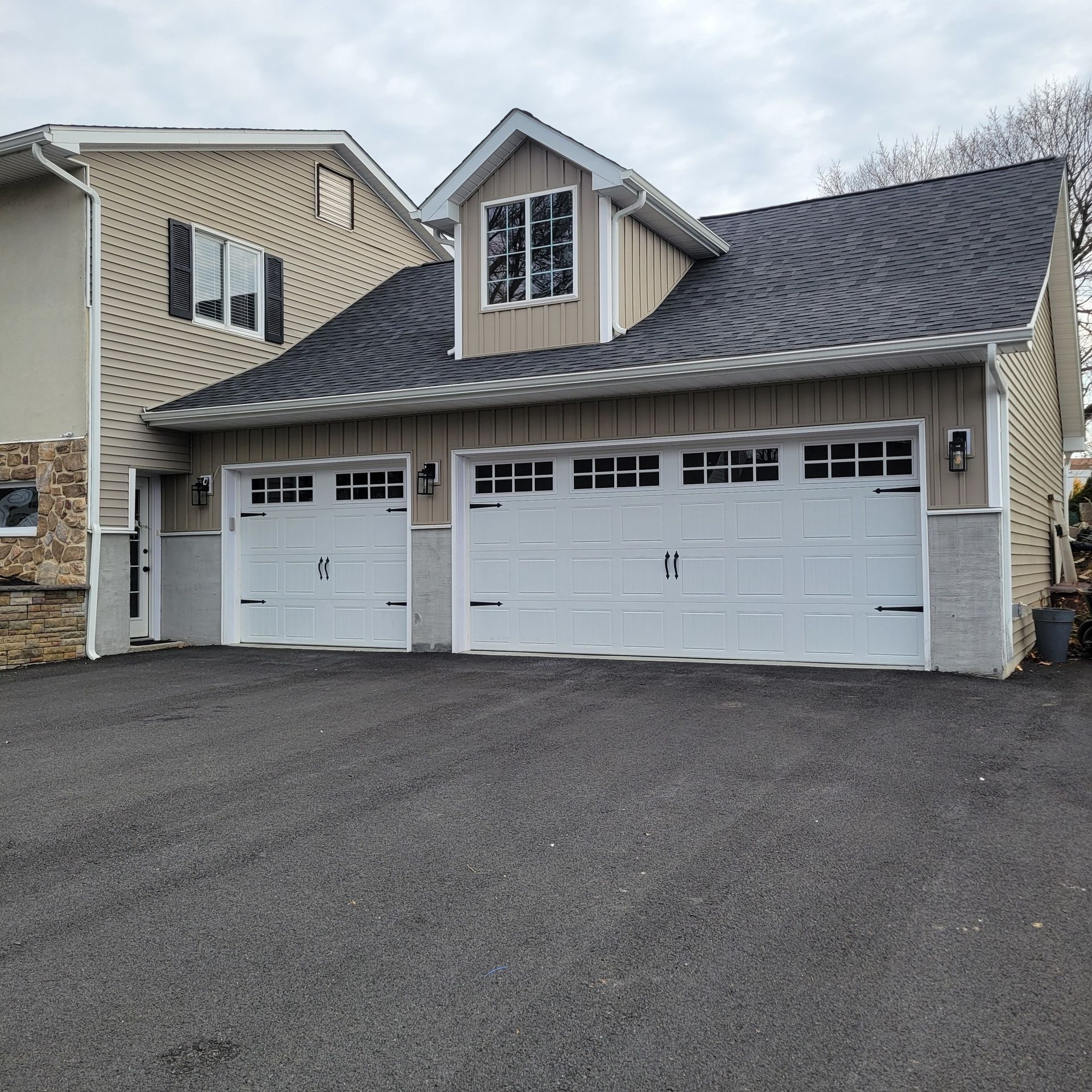 Two-car garage with white doors, tan siding, black roof, and an attached two-story house.