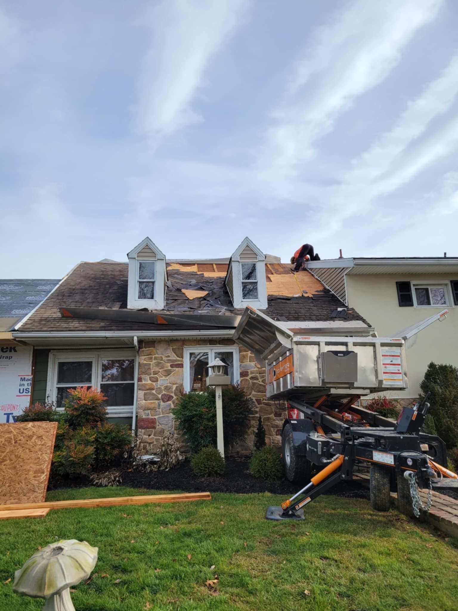 House with damaged roof under repair; worker on roof, lift, and materials on lawn.