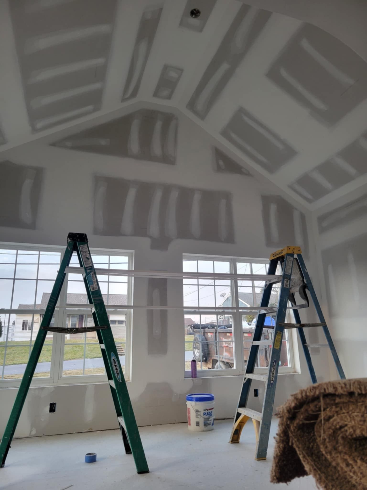 Interior of a house under construction, drywall on walls and ceiling, two ladders, windows, and materials.