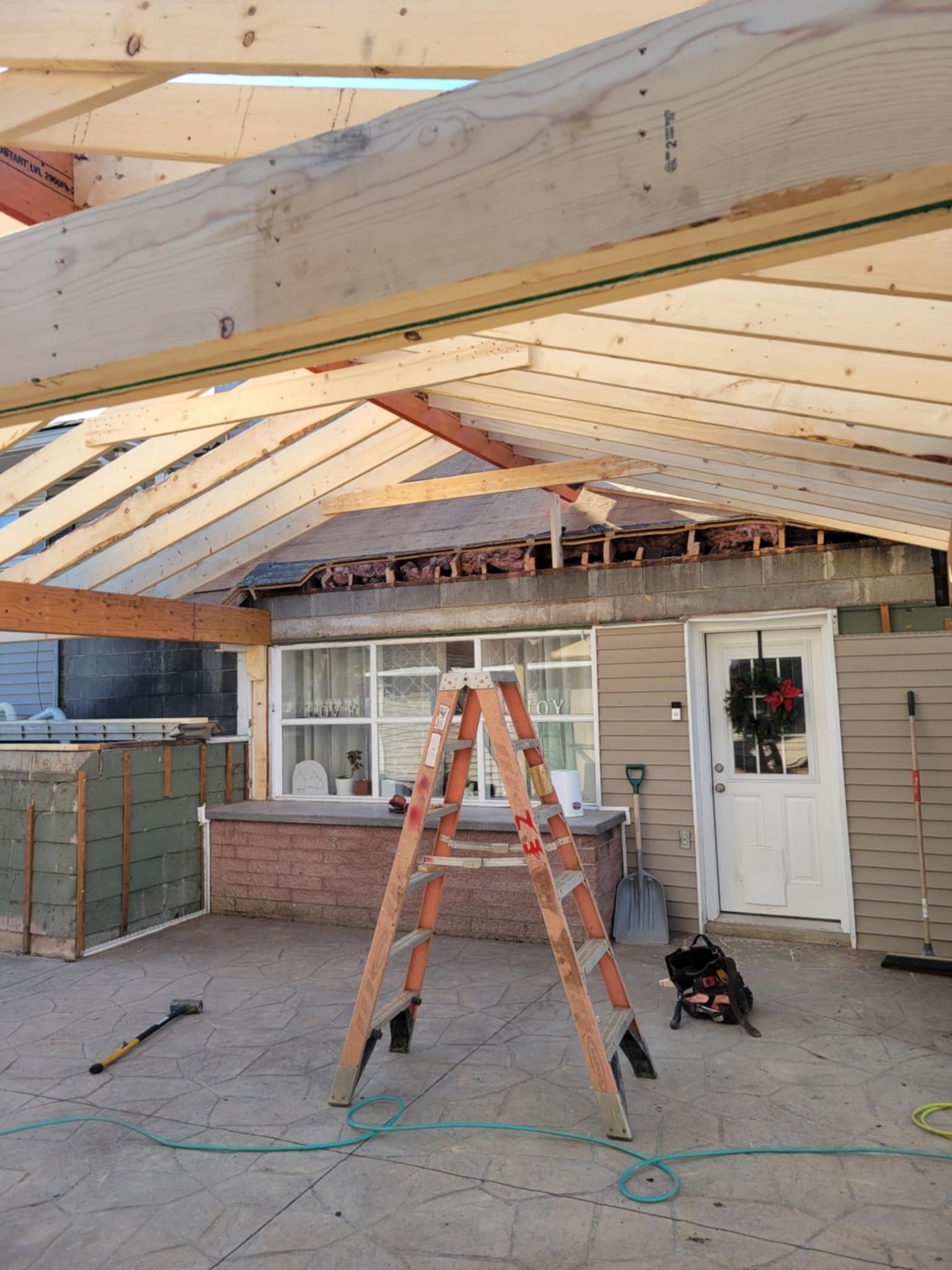 Construction of an outdoor covered patio; ladder in front of house, wood beams, and roofing in progress.