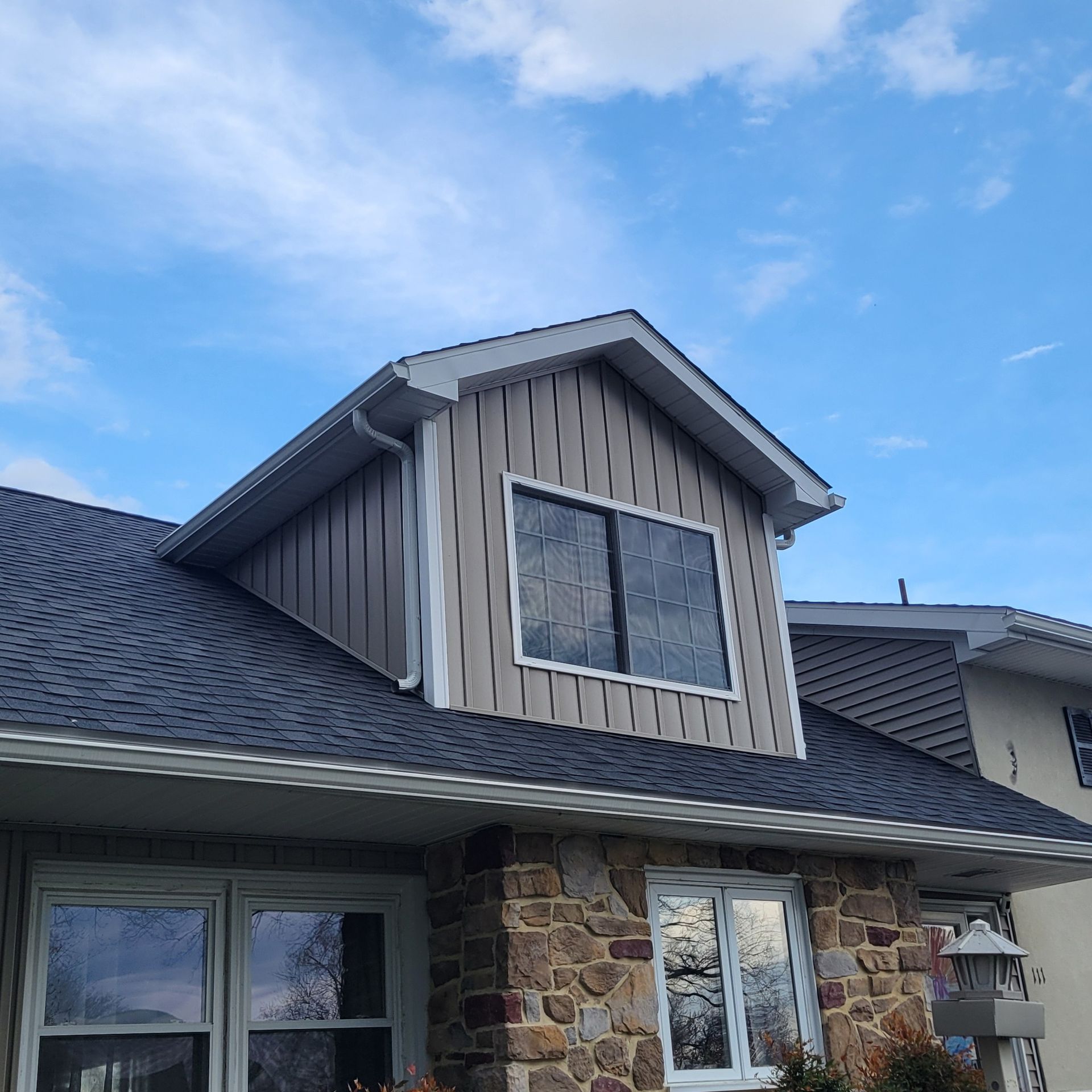Dormer with gray siding and dark roof against a blue sky. Stone and brick house below.