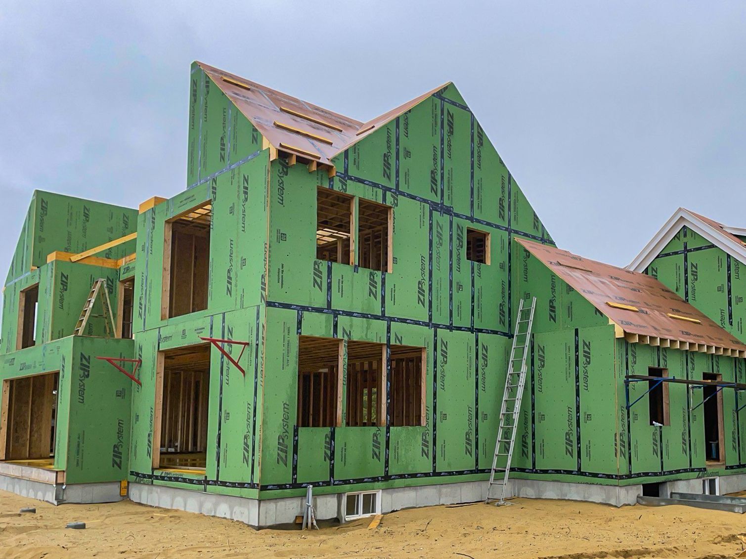 House under construction, green sheathing on wooden frame, cloudy sky, ladder leaning against wall.