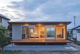 Modern, low-profile house with sliding glass doors opening to a wooden deck. Interior visible, dusk setting.