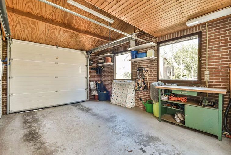Garage interior with garage door open, brick walls, workbench, and windows.