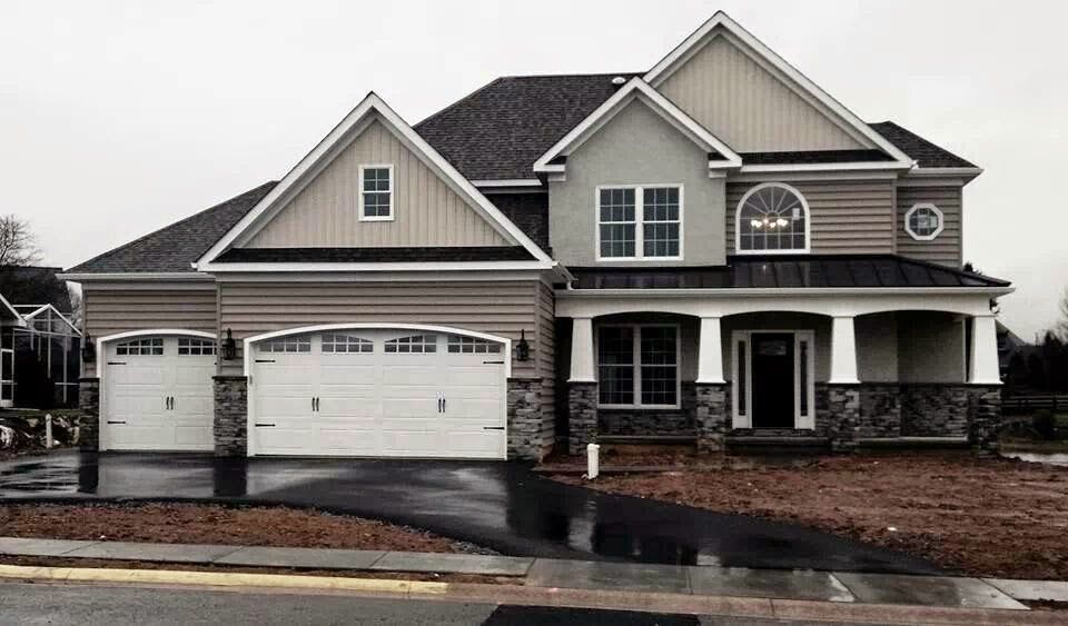 Two-story beige house with a black roof, arched windows, and a driveway. Overcast sky.