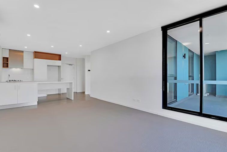 Empty, modern apartment with white walls, kitchen, and a large sliding glass door to a balcony.