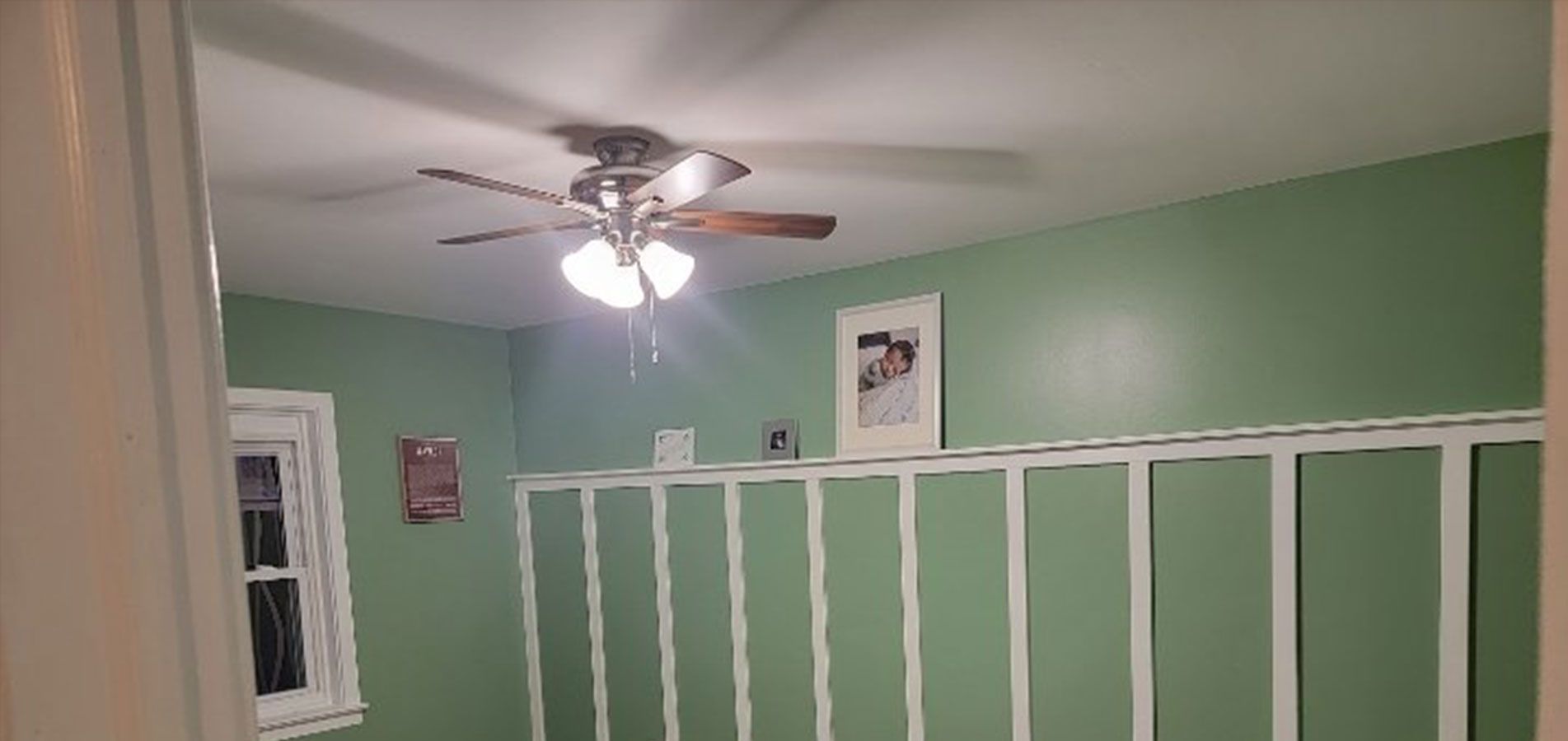 A room with green walls, a ceiling fan, and a white structure. A window and framed photo are visible.