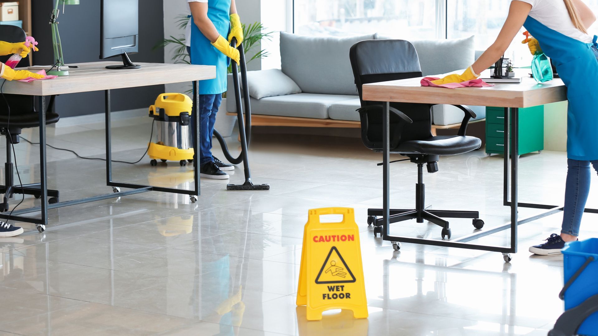 Office cleaners wearing blue uniforms, one vacuuming and another wiping a desk. Wet floor sign in the foreground.
