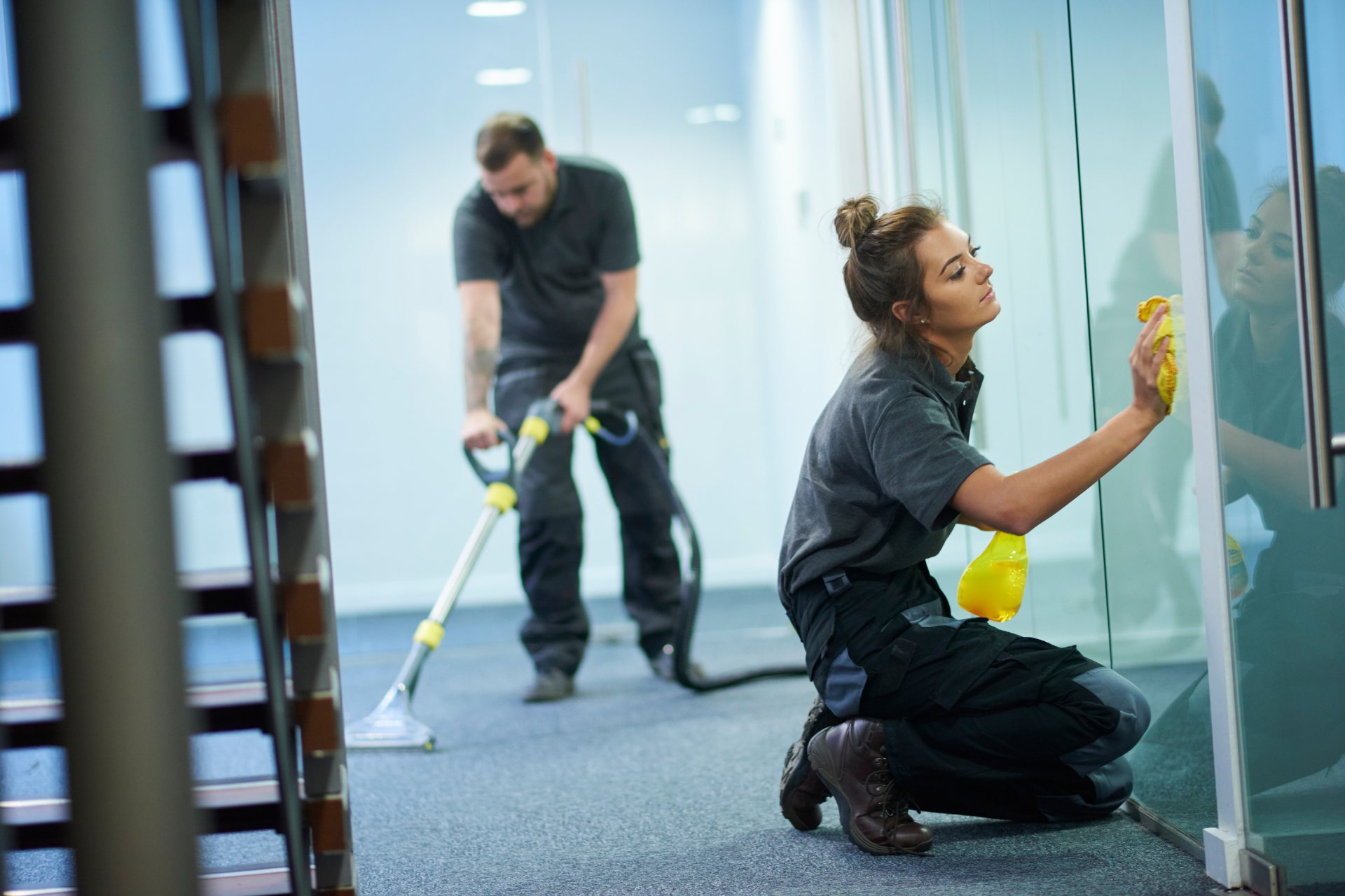 Two cleaning professionals, one wiping glass, the other vacuuming a hallway carpet.