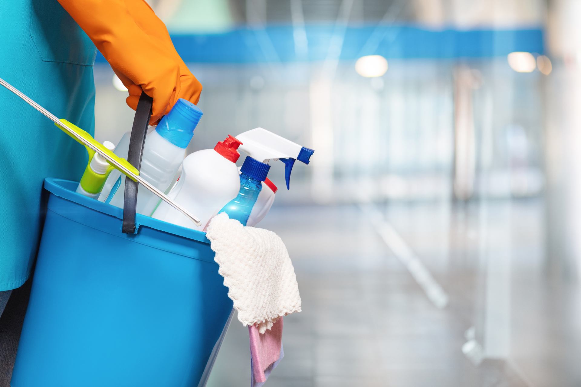 Person in orange gloves holding a blue bucket filled with cleaning supplies in a hallway.
