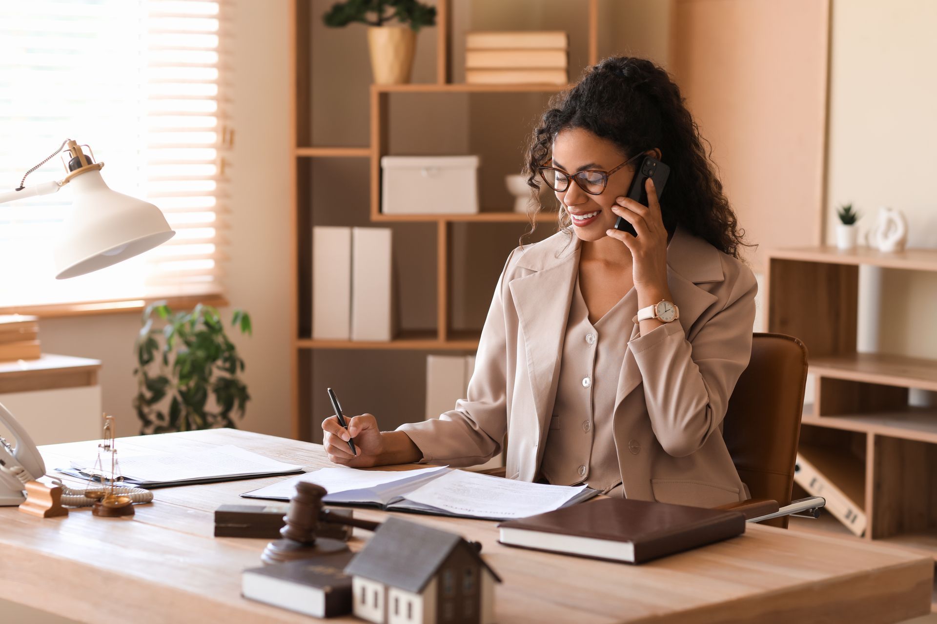 Woman in tan suit on phone, writing at desk in office; documents, gavel, and model house present.