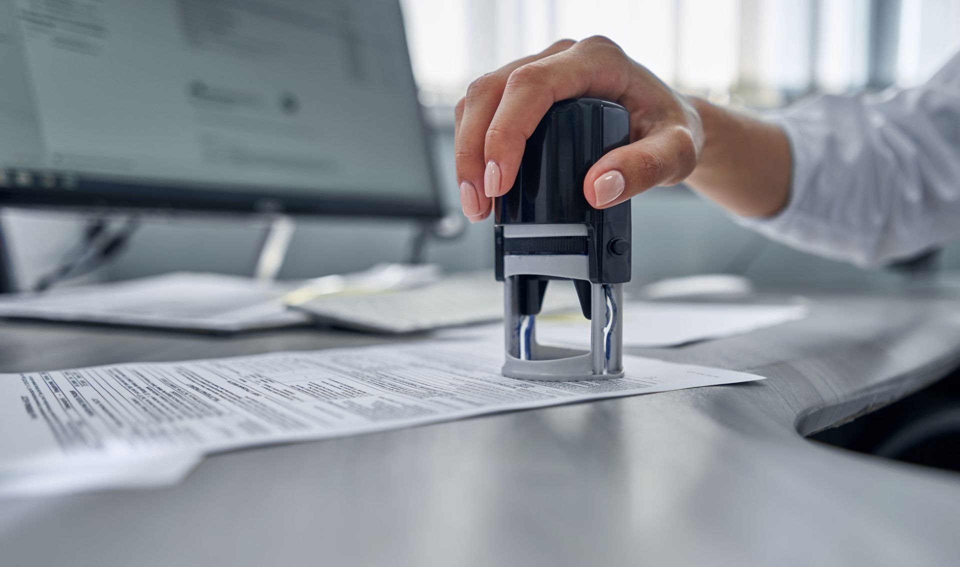 A hand stamps a document with a black office stamp, near a computer and papers on a desk.