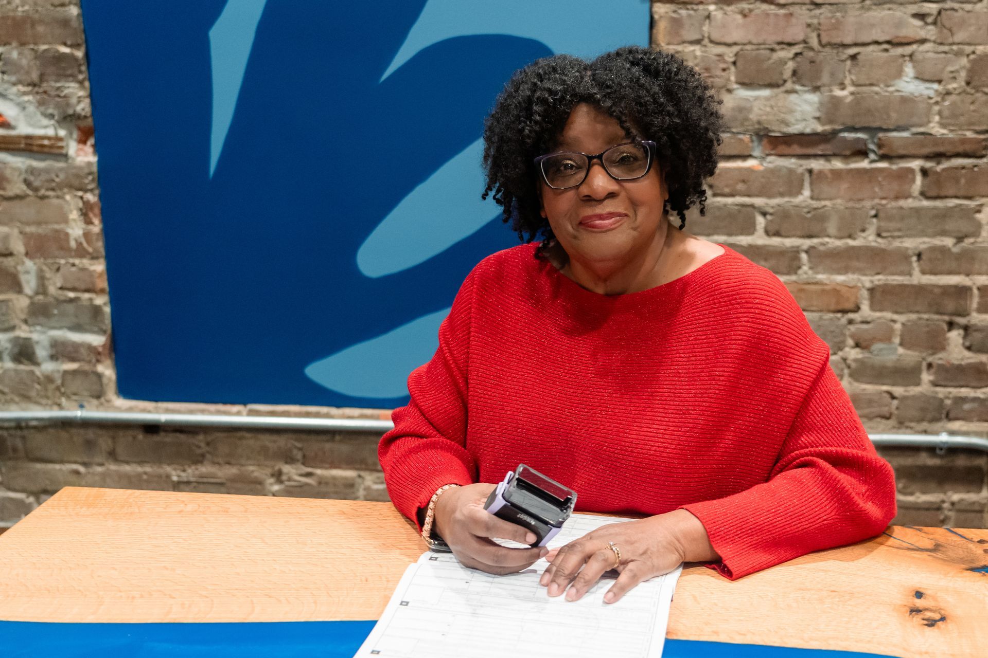 A person in a red sweater sits at a wooden desk with a rubber stamp, working on a document against a brick wall.