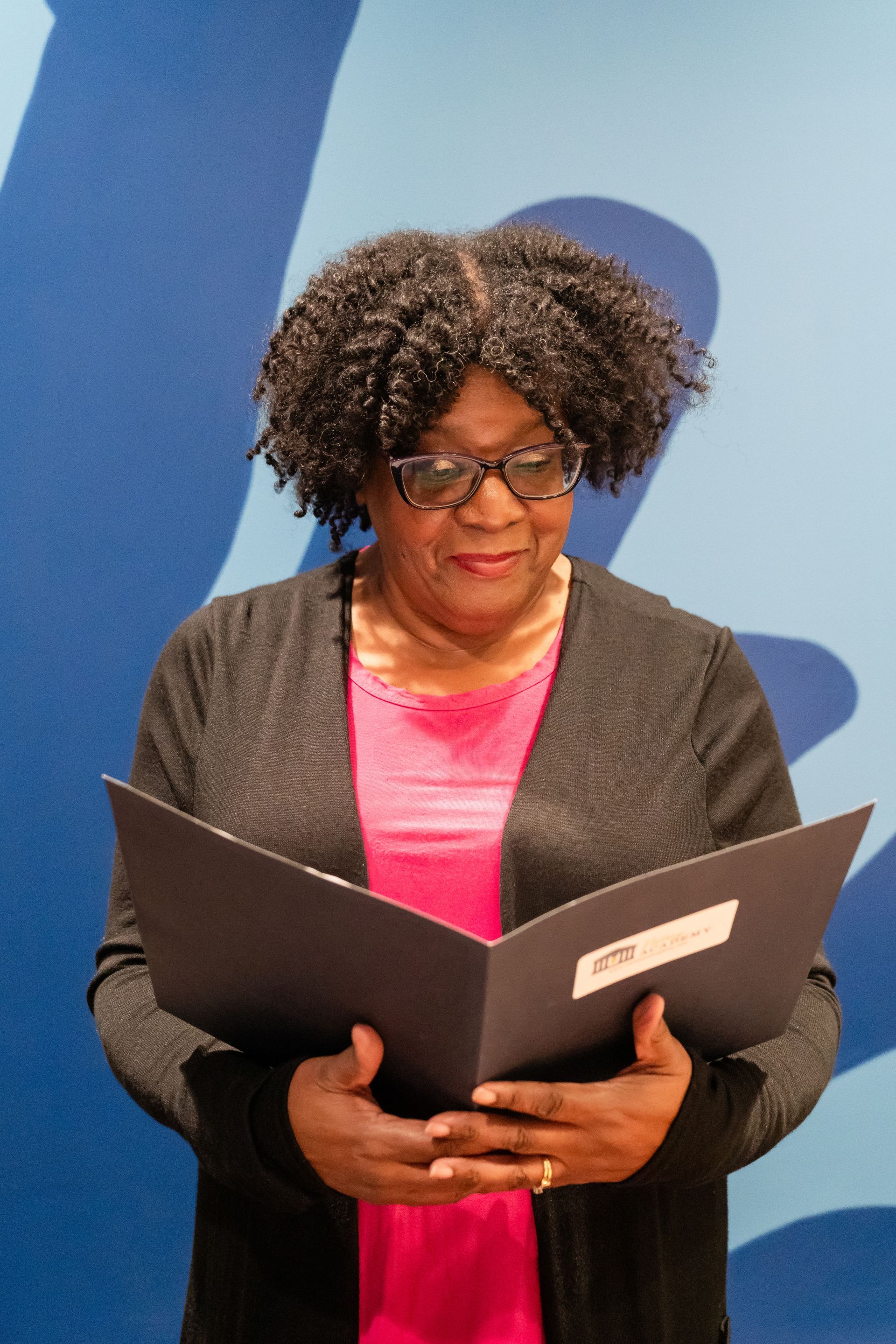 A person wearing a black cardigan and pink top reads from an open black folder against a blue patterned backdrop.