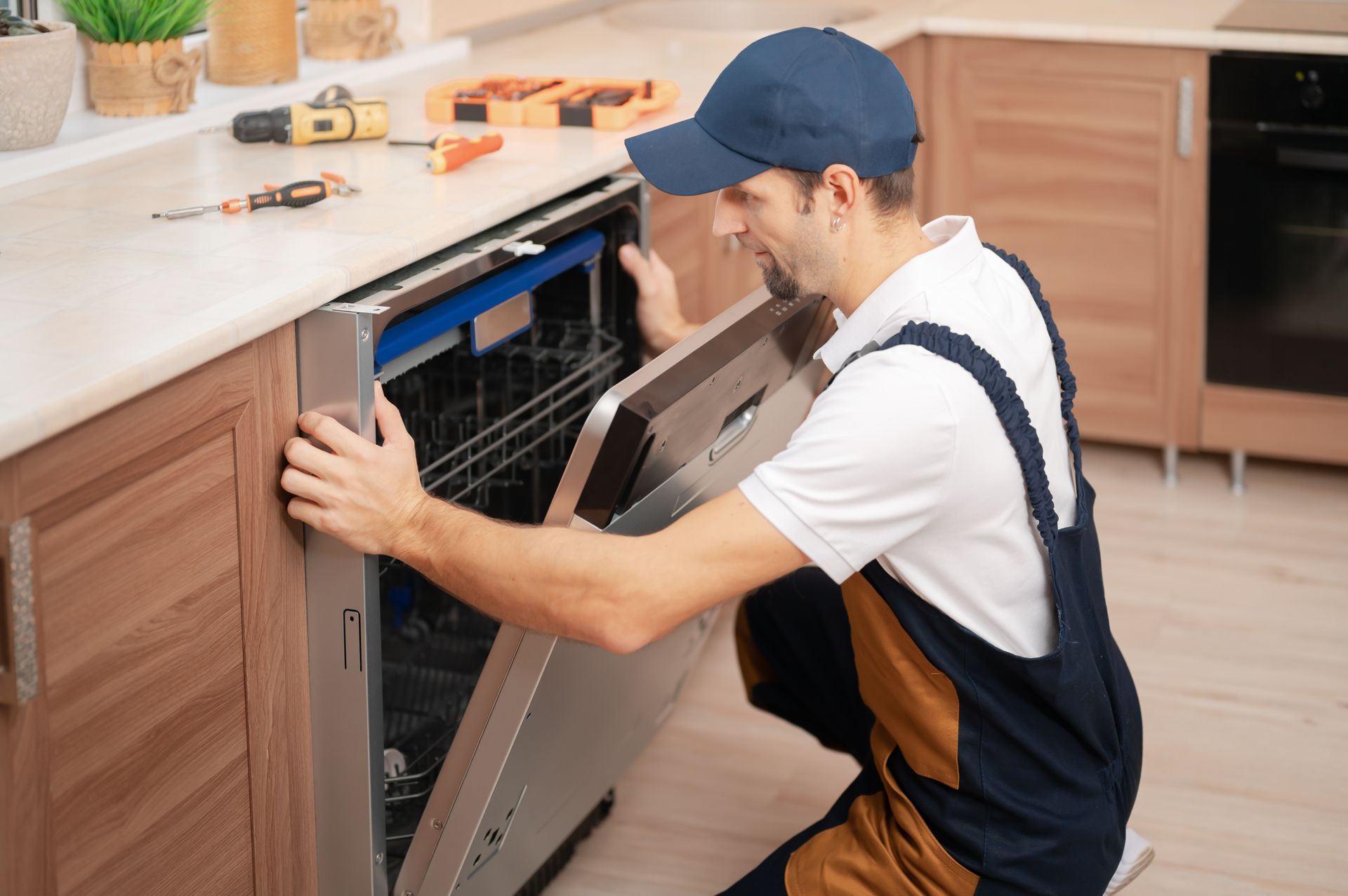 A man in overalls and a cap repairs a dishwasher in a kitchen.