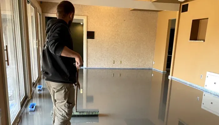 Man applying epoxy floor coating in a room with large windows, using a squeegee.
